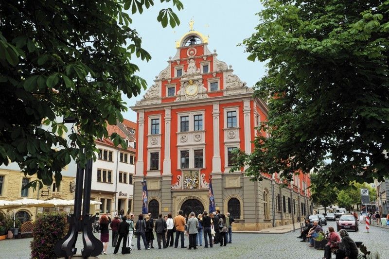 Historisches Gebäude mit roter Fassade und verzierten Giebeln, umgeben von Bäumen und Menschen auf einem Platz.