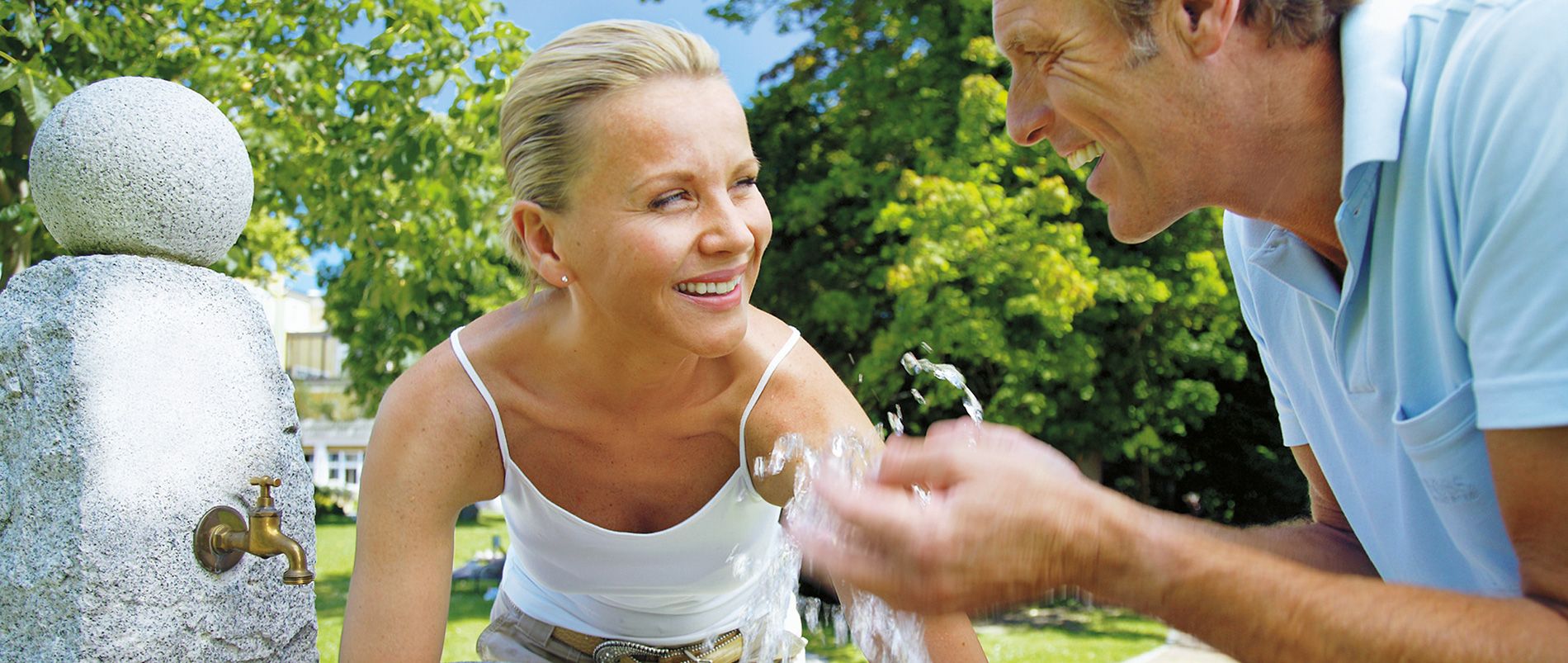 Zwei Personen an einem Brunnen im Freien, lachen und genießen das fließende Wasser.