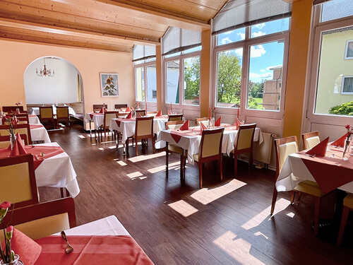 Heller und eleganter Speiseraum mit gedeckten Tischen, roten Servietten und großen Fenstern mit Blick ins Grüne.