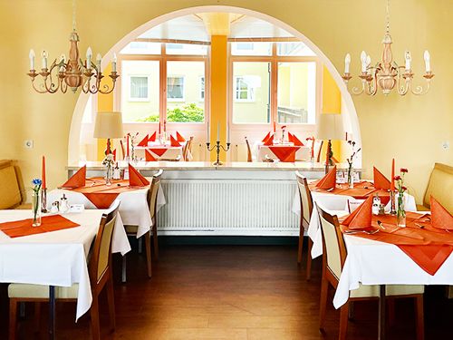 Innenansicht eines elegant gedeckten Restaurants mit weiß gedeckten Tischen und orangefarbenen Servietten, flankiert von Kronleuchtern und großen Fenstern im Hintergrund.
