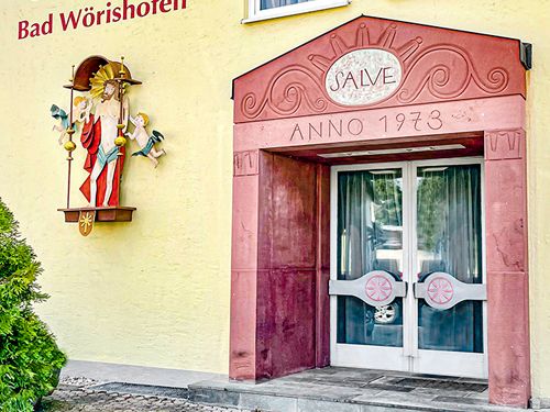 Eingang eines Gebäudes mit der Aufschrift 'SALVE ANNO 1973', flankiert von einem bemalten Wandrelief einer religiösen Figur und dem Schriftzug 'Bad Wörishofen' an der Fassade.