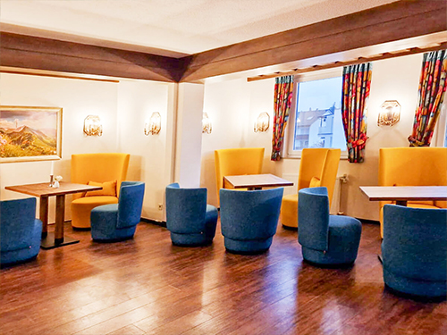 Gemütlicher Raum mit gelben und blauen Sesseln, Holztischen und bunten Vorhängen.