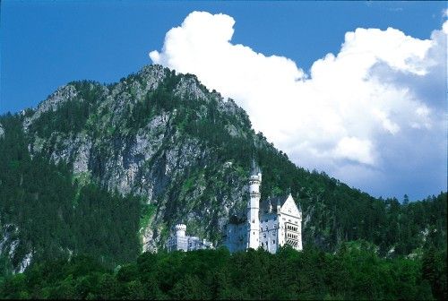 Schloss Neuschwanstein vor einem bewaldeten Berghang unter blauem Himmel mit Wolken.