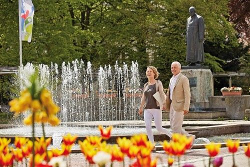 Ein älteres Paar spaziert bei sonnigem Wetter an einem Springbrunnen vorbei, umgeben von blühenden Blumen und einer Statue im Hintergrund.