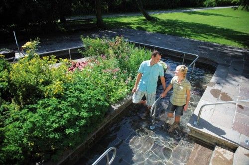 Zwei Personen gehen durch ein Kneippbecken in einem blühenden Garten.
