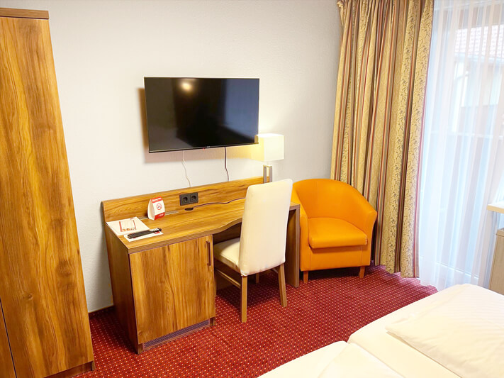 Hotelzimmer mit Holzmöbeln, einem Schreibtisch mit Stuhl, einem Fernseher an der Wand, einem orangefarbenen Sessel und einem Fenster mit Gardinen.