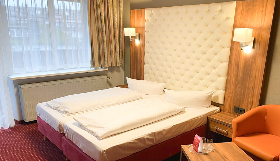 Hotelzimmer mit einem Doppelbett, einem gepolsterten Kopfteil, einer roten Polsterung und einem Holznachttisch mit modernen Lampen.