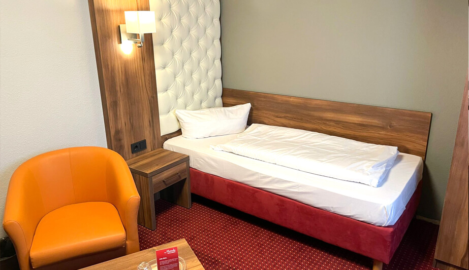 Einzelzimmer mit einem Bett, einem Nachttisch, einer orangefarbenen Sessel und einem rot gepunkteten Teppich.