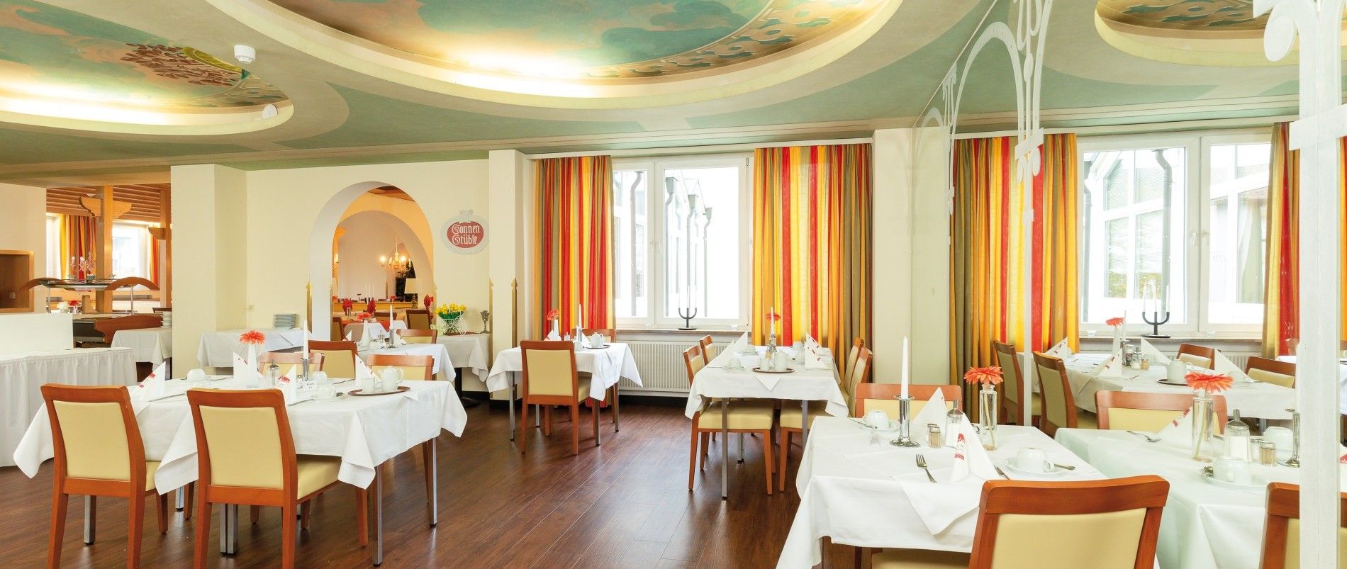 Helles, elegant eingerichtetes Restaurant mit gedeckten Tischen, Holzfußboden und großen Fenstern mit gestreiften Vorhängen.