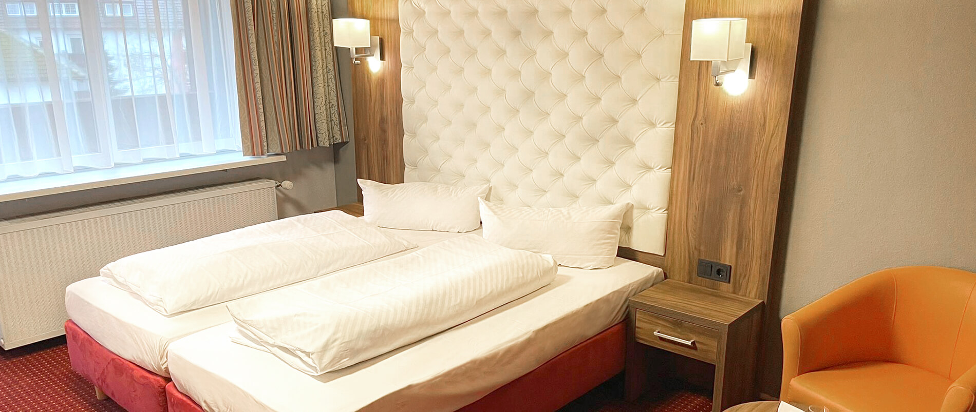 Gemütliches Hotelzimmer mit Doppelbett, gepolstertem Kopfteil, Beistelltisch, Sessel und Fenster mit Vorhängen.
